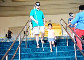 A father, wearing sleepshades, walks down the stairs with a cane. His blind daughter smiles as she leads him down the stairs using her cane.