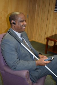 A man smiles as he listens to NFB-NEWSLINE on his iPhone. He sits in a chair with his cane balanced on his shoulder.